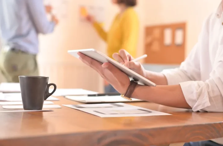Un homme sur sa tablette qui travaille sur son bureau avec deux personnes dans le fond